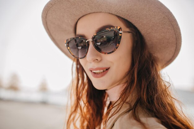 Close-up portrait of fascinating happy woman in hat. Outdoor photo of good-looking girl in dark sunglasses isolated on blur background.