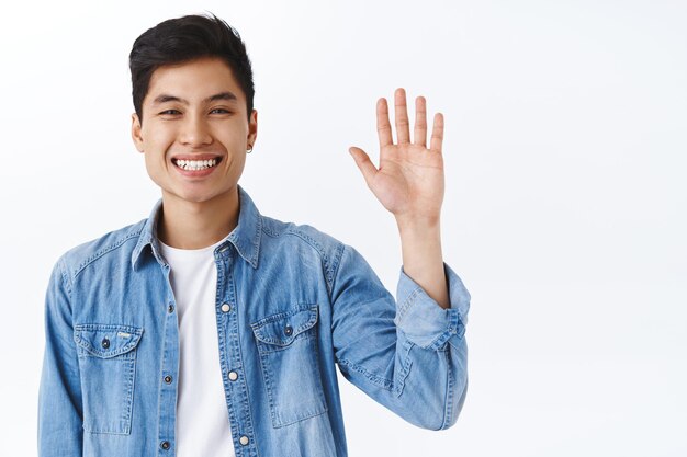 Close-up portrait of friendly attractive asian man waving to say hi, informal greeting gesture, laughing and smiling as welcoming new members join team