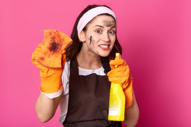 Close up portrait of funny active brunette model points bottle of cleaning detergent at camera, showing dirty orange sponge, having crazy bright facial expression, enjoying time of cleaning up.