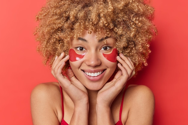 Close up portrait of good looking woman with curly bushy hair touches face gently applies collagen patches for skin treatment wears t shirt shows bare shoulders isolated over red background.