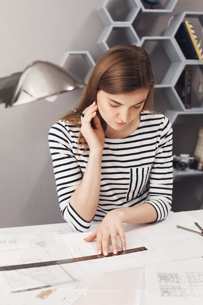 Free photo close up portrait of good-looking young female architect freelancer with dark long hair in striped shirt sitting at white table in coworking space, looking through blueprints she already made, thinkin