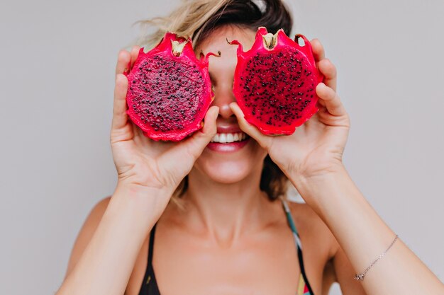 Close-up portrait of good-looking young woman fooling around during photoshoot with dragon fruits. magnificent short-haired girl holding red pitaya.