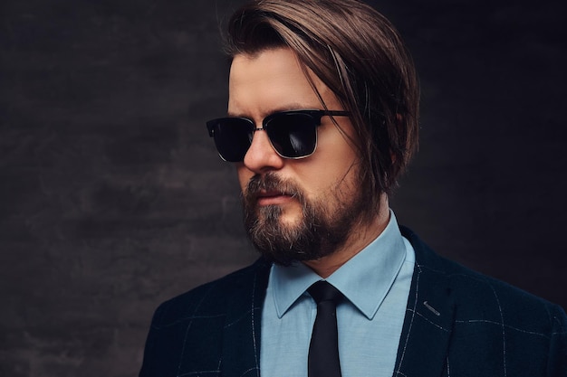 Free photo close-up portrait of a handsome fashionable middle-aged man with beard and hairstyle dressed in an elegant formal suit and sunglasses on a textured dark background in studio.