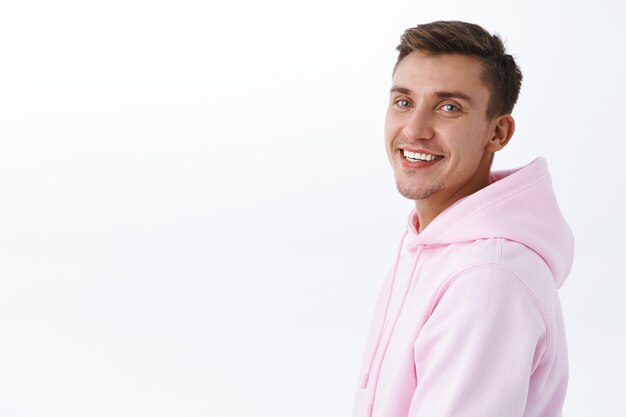 Close-up portrait of handsome young male student with blond hair, bristle, standing in profile and turn to camera with beaming smile, looking satisfied happy, standing white wall