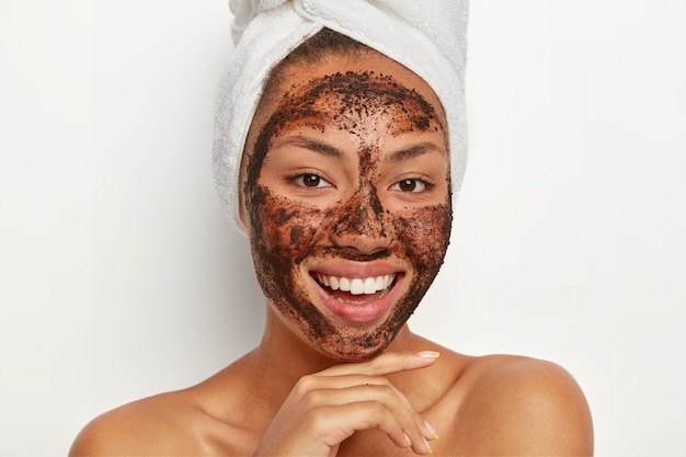 Close up portrait of happy Afro woman touches chin gently, smiles broadly, shows white teeth, cleans face, applies coffee scrub mask, wears wrapped towel on wet hair after taking bath. Skin care