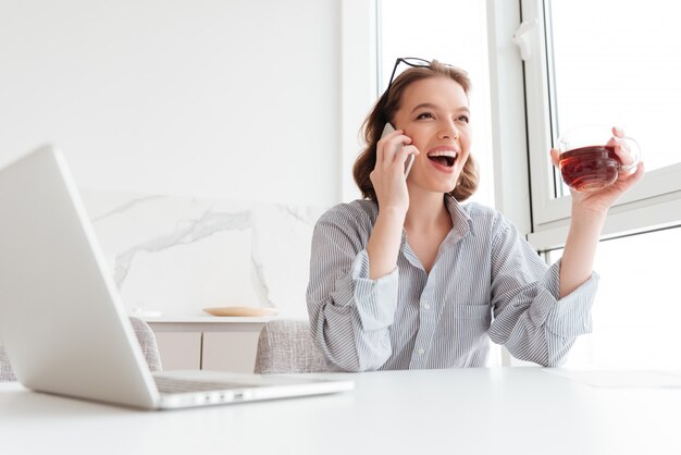 Close-up portrait of happy brunette woman in striped shirt speaking on mobile phone while holding cup of tea, indoors