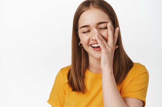 Free Photo close up portrait of happy candid girl, smiling and laughing, touching facial skin with hand, concept of skin care and cosmetic treatment advertising, white background