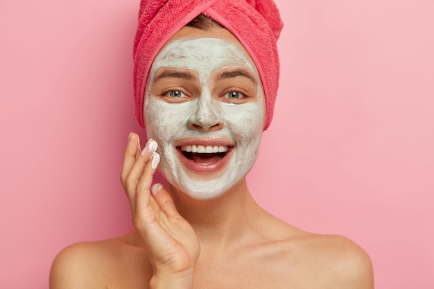 Close up portrait of happy overjoyed female model with cosmetic facial mask applied over her face, has beauty treatments, wears wrapped towel on head, has healthy refreshed look. Renewal and therapy