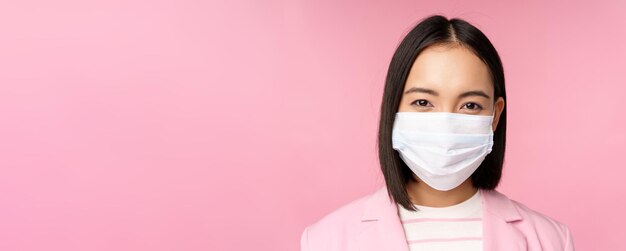 Close up portrait of japanese businesswoman in medical face mask suit looking at camera standing over pink background