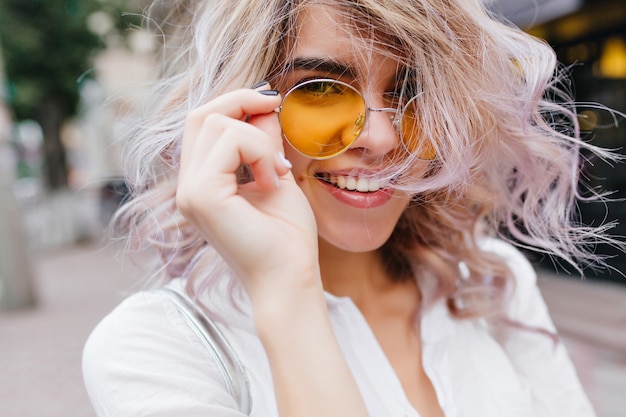 Close-up portrait of joyful fair-haired lady spending time outside holding trendy yellow glasses