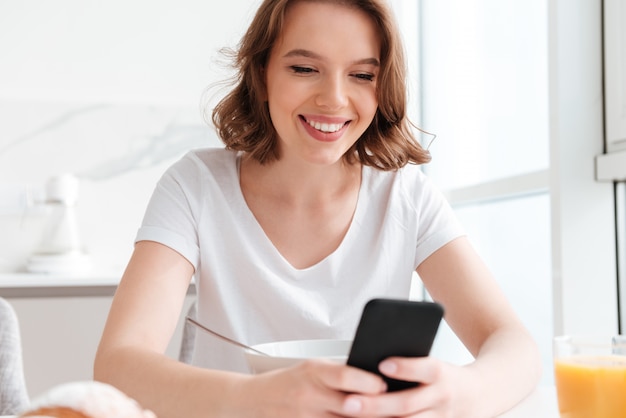 Free photo close-up portrait of joyful woman texting message on smartphone while sitting and having breakfast at the kitchen