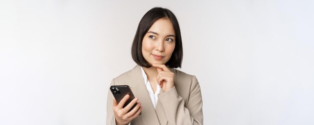 Close up portrait of korean woman corporate lady in suit using mobile phone and smiling holding smartphone standing over white background