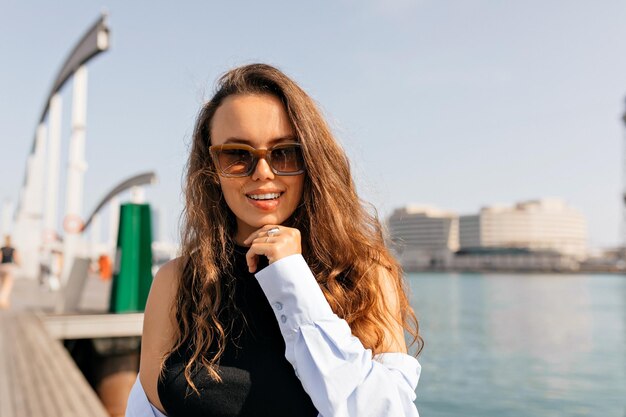 Close up portrait of longhaired european model in sunglasses and blue shirt posing at camera on background of lake Young attractive caucasian woman laughing full mouth swinging hair outdoors