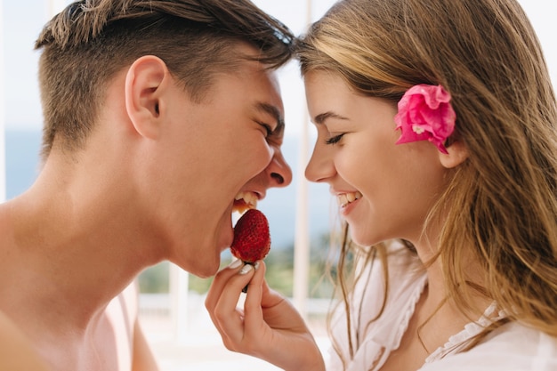 Free photo close-up portrait of loving young couple posing with eyes closed, while eating strawberry. adorable blonde girl with pink flower in hair feeds her boyfriend with tasty berries.