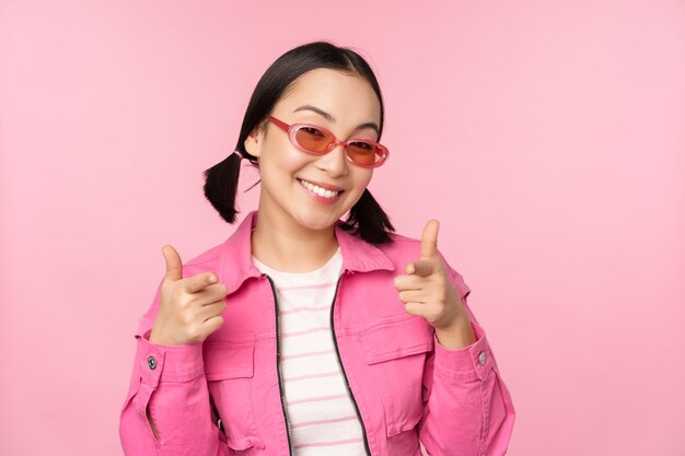 Close up portrait of modern asian girl in sunglasses smiling pointing fingers at camera praise you inviting or complimenting standing over pink background