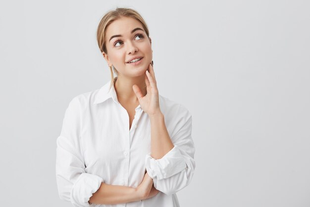 Close-up portrait of pleased woman with pure skin, dark eyes and sincere smile wearing white shirt posing. Dreamy woman thinking about pleasant things, looking upwards.