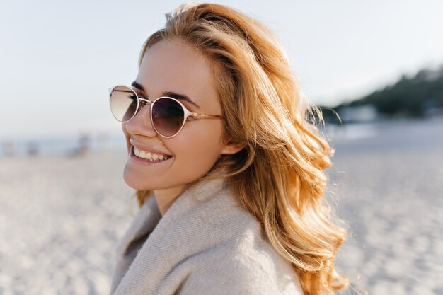 Close-up portrait of positive woman with wavy blinde hair dressed in beige cashmere sweater and sunglasses in beach.