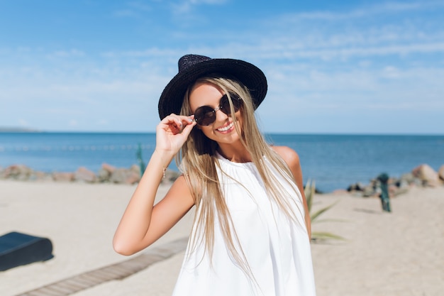 Free photo close-up portrait of pretty blond girl with long hair is standing on the beach near the sea. she is  smiling to the camera.