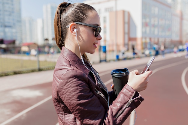 Free Photo close-up portrait of pretty lady with dark collected hair wears black sunglasses drinks coffee, looking in the phone and listening to music in earphones