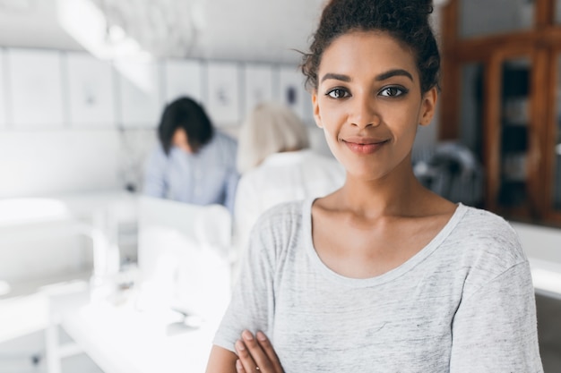 Free photo close-up portrait of pretty mulatto woman with trendy make-up standing with arms crossed in office. indoor photo of female black employee posing with international colleagues behind and gently smile.