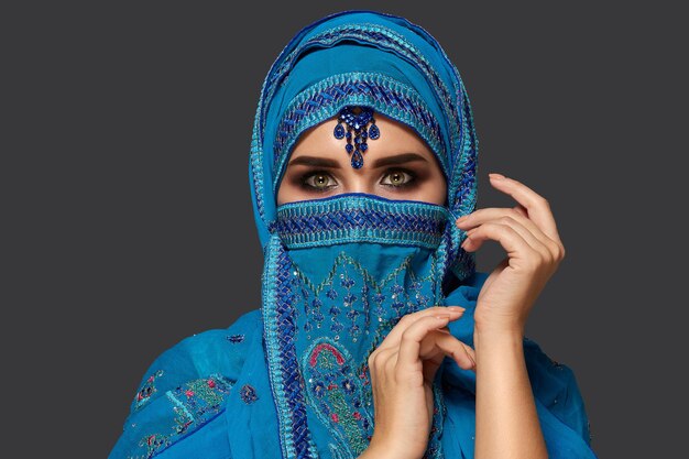 Close-up portrait of a pretty woman with beautiful smoky eyes wearing a blue hijab decorated with sequins and jewelry. She is gesticulating and looking at the camera on a dark background. Human emotio