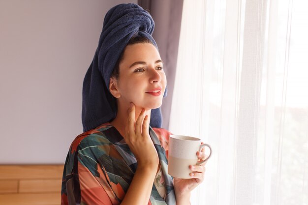 Close up portrait of pretty woman wrapped in towel and bathrobe waking up in morning sitting on bed and drinking coffee