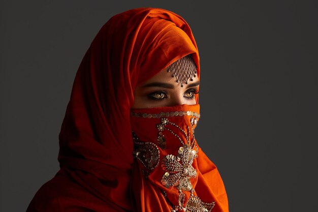 Close-up portrait of a pretty young female with beautiful smoky eyes and jewelry on the forehead, wearing the terracotta hijab decorated with sequins. She is standing sideways and looking away on a da