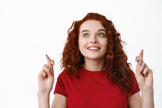 Free Photo close up portrait of redhead hopeful woman with curly hair cross fingers for good luck staring at upper left corner logo with tempted face wants something praying or making wish