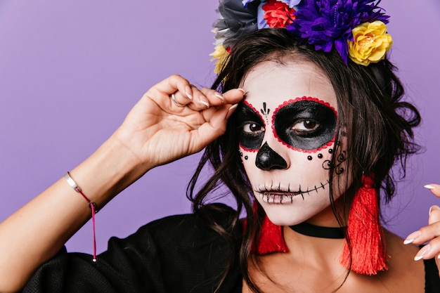 Close-up portrait of refined woman with dark eyes posing in masquerade costume. Cute latin lady in flower wreath preparing for halloween.