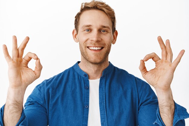 Close up portrait of satisfied redhead man, smiling and looking happy, showing OK okay gesture to praise something good, well done great gesture, recommending excellent service, white wall