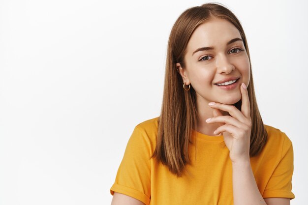 Close up portrait of smiling blond woman look intrigued, smiling thoughtful, interesting suggestion, standing pleased in yellow t-shirt against white wall