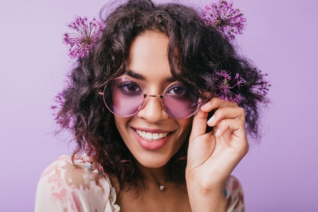 Close-up portrait of smiling brown-eyed woman with flowers in black hair. African blissful lady in sunglasses posing.