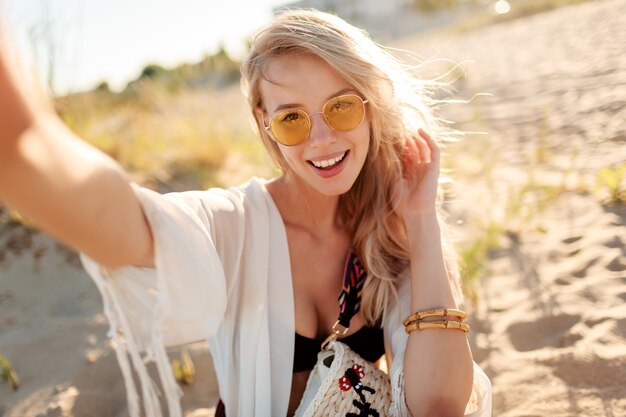 Free photo close up portrait of smiling carefree woman with blonde hairs making self portrait on sunny evening beach.