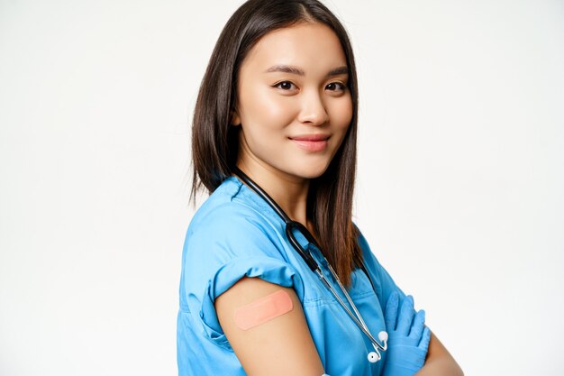 Close up portrait of smiling, confident female doctor in scrubs, showing arm with patch after covid-19 vaccination, vaccinated healthcare worker, standing over white background.