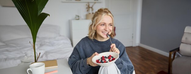 Close up portrait of smiling cute blond woman eating healthy lunch in her room holding bowl with