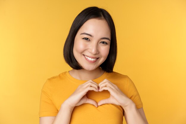 Close up portrait of smiling korean woman showing romantic heart sign and looking happy standing over yellow background Copy space