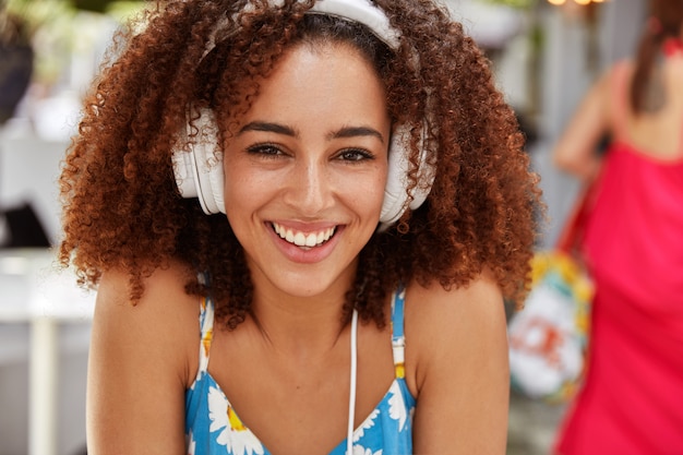 Close up portrait of smiling satisfied beautiful female with dark skin, has crisp hair, enjoys spare time during summer holidays in sidewalk cafe