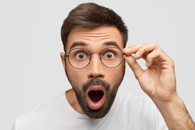 Close up portrait of stunned bearded young guy drops jaw, has bugged dark eyes, sees something unbelievable and surprising, has eyewear, isolated on white wall. People, emotions concept