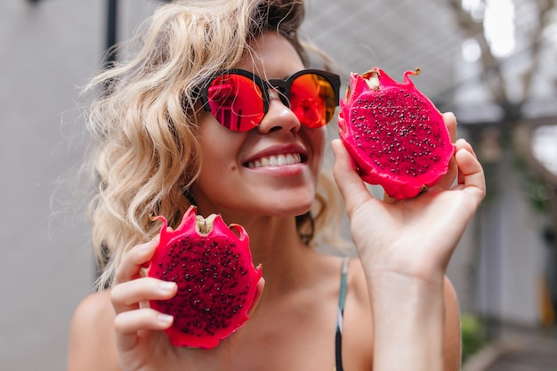 Free photo close-up portrait of stunning blonde girl in pink sunglasses posing with exotic fruits. photo of laughing curly female model with red pitahaya.