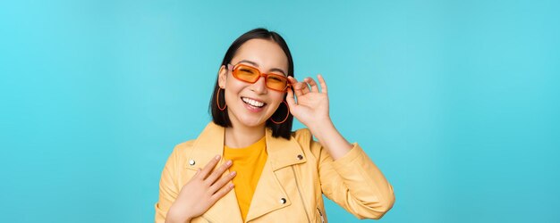 Close up portrait of stylish asian woman in sunglasses laughing and smiling looking happy posing in trendy clothes over blue background