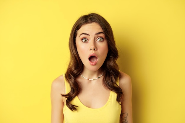 Close up portrait of stylish brunette girl looking amazed, wow face, showing excitement and surprise, impressed by something, standing over yellow background