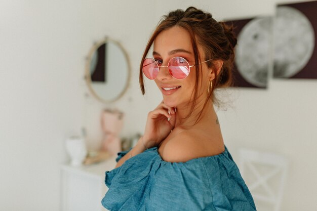 Close up portrait of stylish charming woman with wonderful smile wearing round glasses and blue tshirt smiling at camera over light background