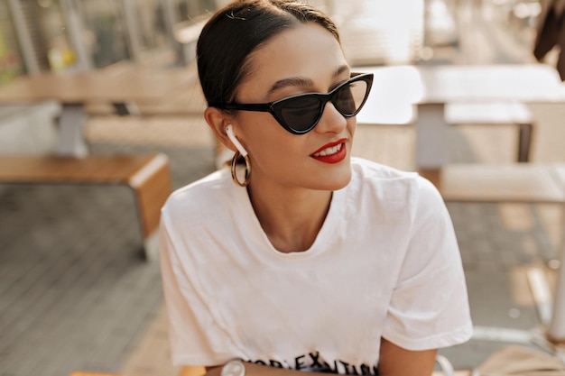 Close up portrait of stylish European woman with dark hair and red lips wearing glasses and white shirt is posing at camera while resting outdoor