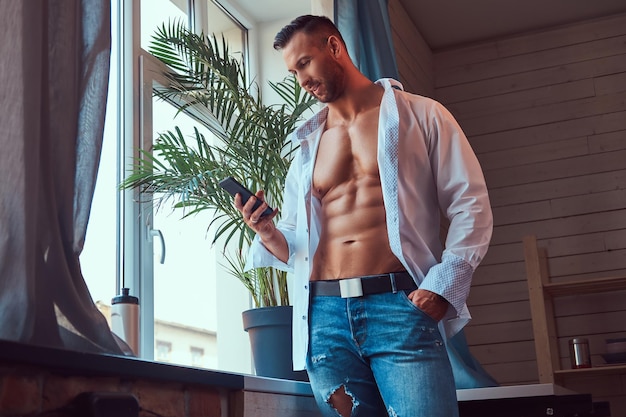 Close-up portrait of a tall bearded male with a muscular body, dressed in a white shirt and jeans, using a smartphone.