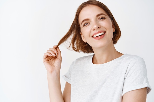 Close-up portrait of tender girl with short hair, smiling white teeth and looking happy, playing with haircut, standing against white wall