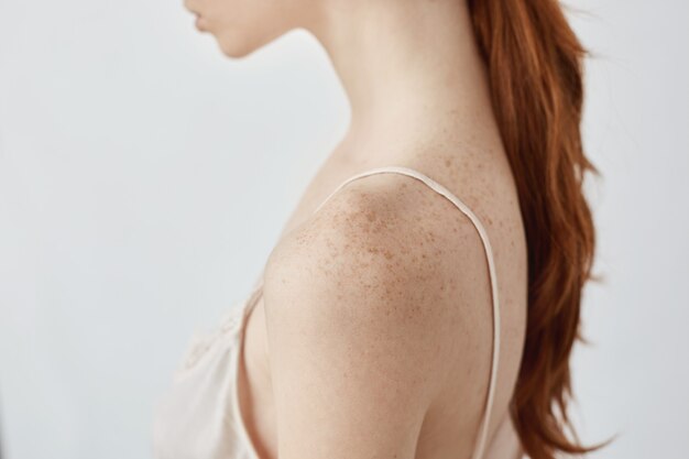 Close up portrait of tender young redhead freckled girl in lingerie over white wall.