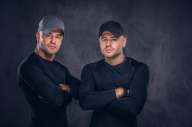 Free photo close-up, a portrait of two handsome young male dressed in a black sportswear and baseball cap over a dark background.