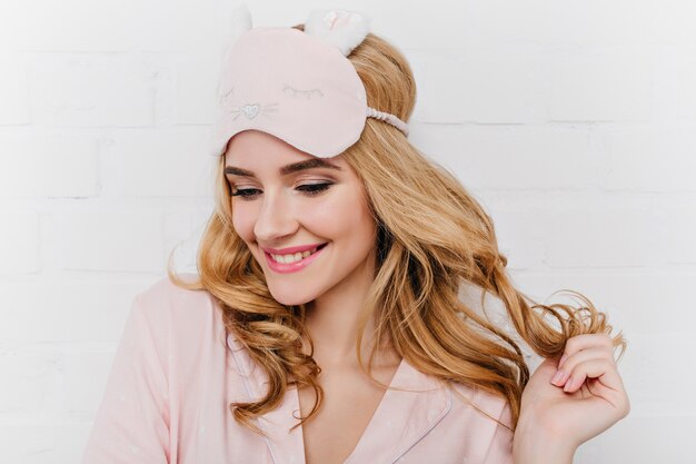 Close-up portrait of woman with glamorous hairstyle posing in weekend morning. Joyful white girl in sleep mask playing with curly hair.