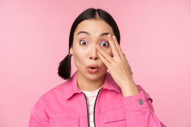 Close up portrait of young asian girl looking surprised express amazement and wonder peeking through fingers standing over pink background