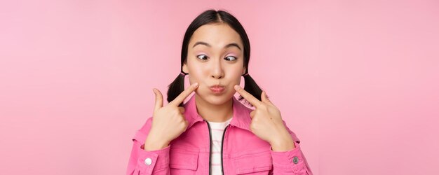 Close up portrait of young asian girl showing her dimples poking cheeks silly and making funny faces standing over pink background